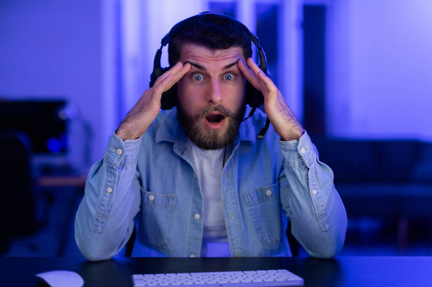 Shocked man gamer with hands on head reacting to an unbelievable moment in a game at night, home interior - Photo, Image