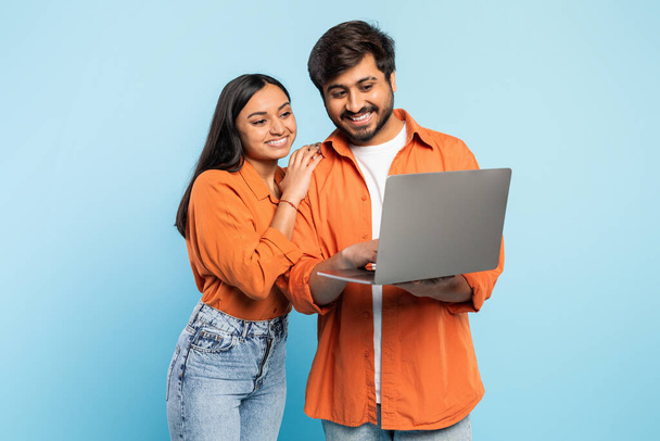 Индийская пара в оранжевой одежде улыбается, сотрудничая и глядя на экран ноутбука, современная концепция работы - Фото, изображение