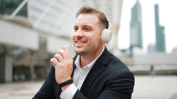 Ammattimainen liikemies kuuntelee kuulokkeita ja käyttää puhelinta äänimerkintä istuessaan portaikossa kaupungissa. Manageri käyttää kuulokkeita kuunnellen rentoa laulua ja liikkuen vilkkaalla tuulella. Kaupungit. - Valokuva, kuva