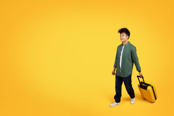 Молодий чорний чоловік ходить, тримаючи жовту валізу, дивлячись на простір для копіювання з цікавістю на жовтому тлі - Фото, зображення