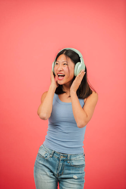 Verticaal. Jonge Aziatische vrouw dansende muziek met behulp van een draadloze hoofdtelefoon en een streaming mobiele telefoon app op rode achtergrond. Chinese tiener vrouw luisteren naar een lied met oortjes op roze kleur achtergrond muur - Foto, afbeelding