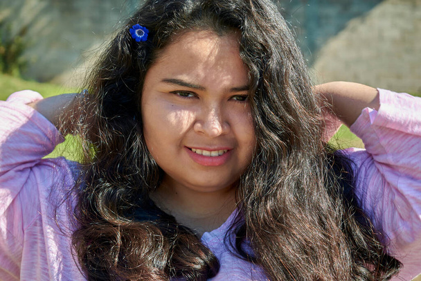 Portret młodej latynoskiej kobiety w letni dzień. Ma długie czarne włosy. Uśmiecha się do kamery. Ma niebieski kwiat we włosach.. - Zdjęcie, obraz