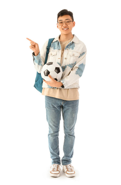 Feliz estudiante asiático masculino con mochila y pelota de fútbol apuntando a algo sobre fondo blanco - Foto, Imagen