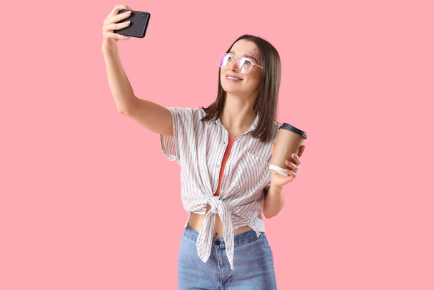 Mladá žena v brýlích s šálkem kávy, přičemž selfie na růžovém pozadí - Fotografie, Obrázek
