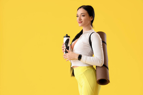 Portrait of sporty young woman with water bottle and yoga mat on yellow background - Фото, изображение