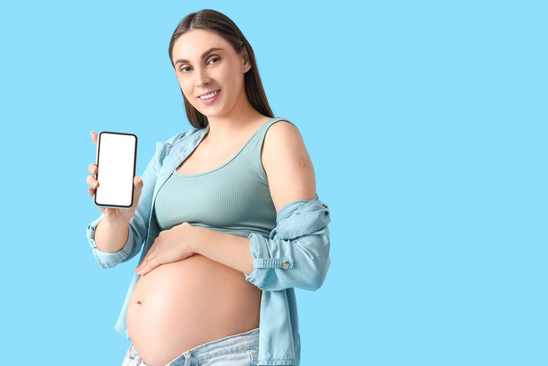 青い背景でのワクチン接種後の携帯電話と医療パッチを持つ若い妊婦 - 写真・画像