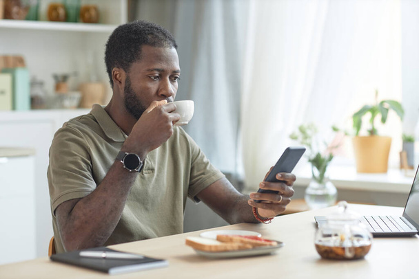 Portret dorosłego Afroamerykanina korzystającego ze smartfona podczas jedzenia śniadania przy stole kuchennym z laptopem - Zdjęcie, obraz