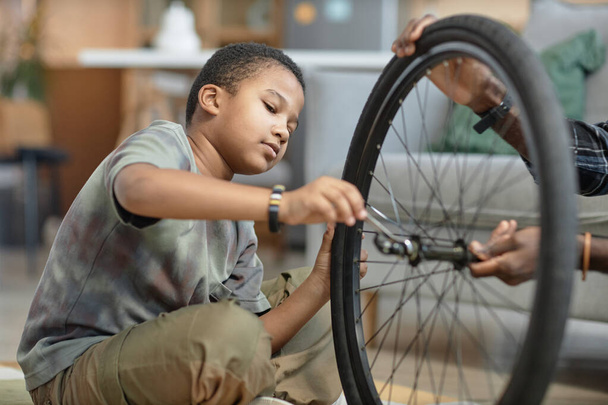 自転車の車輪を修理する若いブラックボーイのサイドビュー肖像画,父親が自宅で床に座っている - 写真・画像