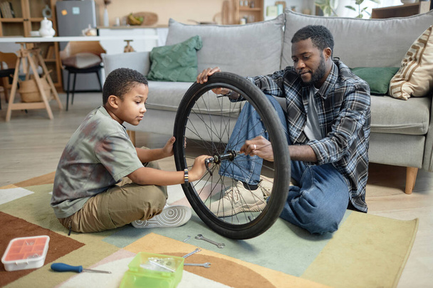 Vista laterale ritratto di Nero giovane padre e figlio fissaggio ruote bicicletta insieme a casa - Foto, immagini