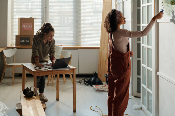ワークウェアの若いアフリカ系アメリカ人女性は,ノートパソコンでテーブルの上に曲がっている同僚に対して立っている間,ペイントローラーで壁を描きます - 写真・画像