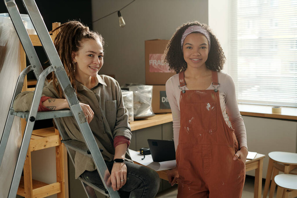 Δύο χαρούμενες νεαρές σχεδιάστριες εσωτερικών χώρων ή ζωγράφοι με ενδύματα εργασίας κοιτάζοντας την κάμερα, ενώ η γυναίκα με dreadlocks κάθεται στη σκάλα - Φωτογραφία, εικόνα