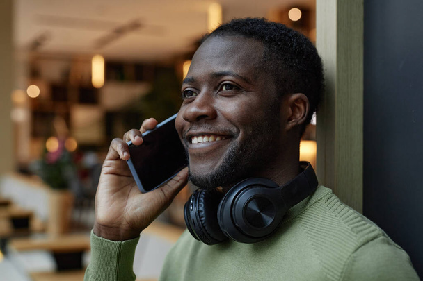 Side view portrait of Black young man speaking on phone and smiling in modern office lounge copy space - Photo, Image