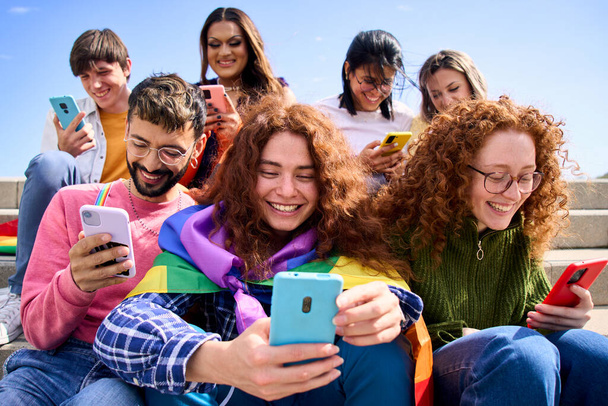 Uśmiechnięci młodzi przyjaciele korzystający z telefonu komórkowego świętujący wspólnie festiwal gejów. Szczęśliwa społeczność LGBT z tęczowymi flagami oglądająca i sprawdzająca portale społecznościowe na zewnątrz - Zdjęcie, obraz