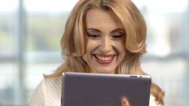 Portrait d'une heureuse femme caucasienne blonde d'âge moyen utilisant une tablette numérique à l'intérieur. Fenêtre intérieure en arrière-plan. - Séquence, vidéo