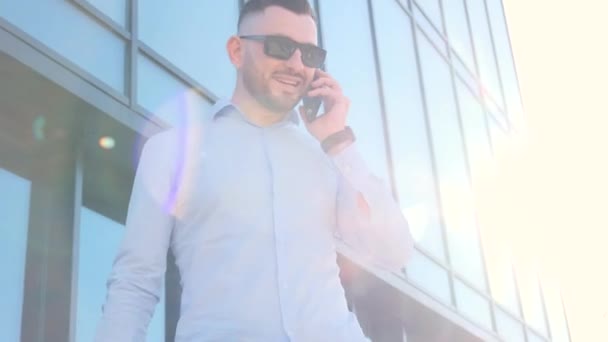 Vonzó fiatal üzletember beszél telefonon a napfény hátterében. A cég igazgatója az üzleti központ üvegépületének hátterében - Felvétel, videó