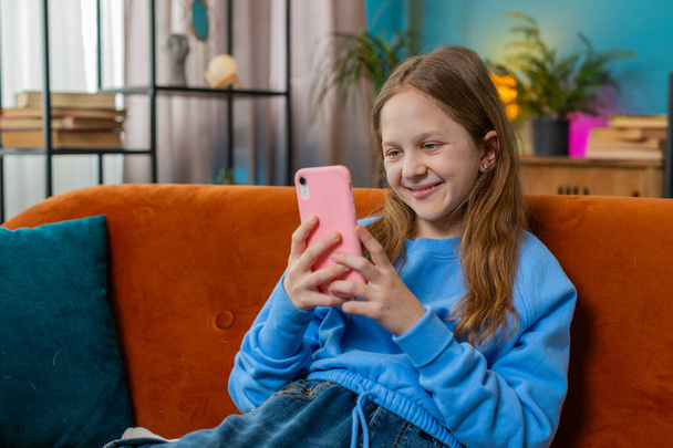 Fröhliche preteen Mädchen SMS teilen Nachrichten auf Smartphone Social-Media-Anwendungen online anschauen Relax-Film. Weibliche kaukasische Teenager-Kind mit Handy zu Hause im Wohnzimmer sitzt auf Sofa. - Foto, Bild