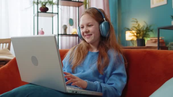 Biała nastolatka w słuchawkach z laptopem słucha muzyki lub lekcji, uczy się na odległość. Edukacja online. Uśmiechnięte dziecko relaksujące się, robiące sobie przerwę w pracy w domu, siedzące na kanapie. - Materiał filmowy, wideo