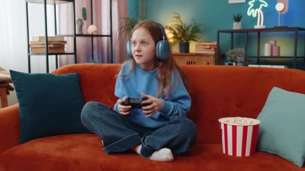 Veselá šťastná mladá dívka dítě ve sluchátkách pomocí ovladače joystick hraje videohry televizní hra zábava těší sedí na pohovce v pokoji. Kavkazský preteen dítě těší online hry doma - Záběry, video