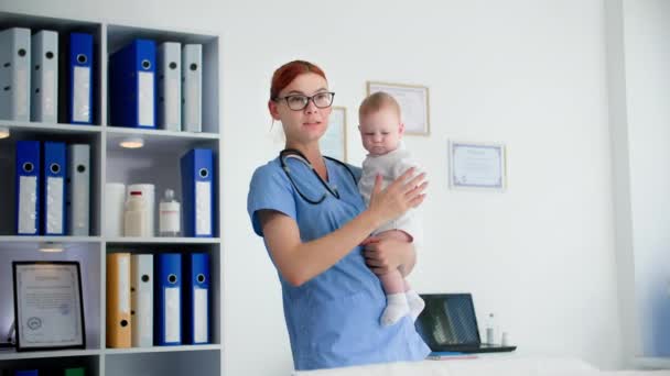 portrait d'une jeune femme en uniforme médical avec des lunettes tenant une petite fille dans les bras dans un cabinet de médecin, souriant et regardant la caméra - Séquence, vidéo