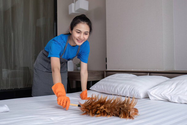 Hotellin sisäkkö univormussa puhdistaa sängyn ja makuuhuoneen höyhenpölyllä.. - Valokuva, kuva
