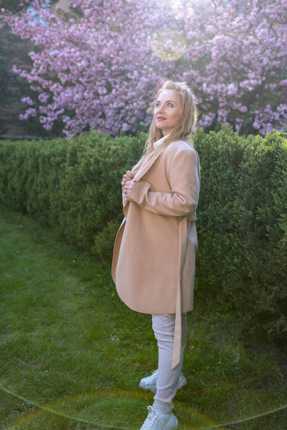Nachdenkliche Frau mittleren Alters in beigem Mantel mit langen blonden Haaren im Frühlingspark. Ein Porträt in voller Länge. Vertikaler Rahmen - Foto, Bild