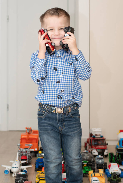 Filhos. Carros de brinquedo. Um menino pequeno, alegre e bonito, de 4 anos, vestindo uma camisa quadriculada, brinca com muitos carros coloridos em um interior de casa. Os brinquedos são colocados uniformemente no chão. - Foto, Imagem