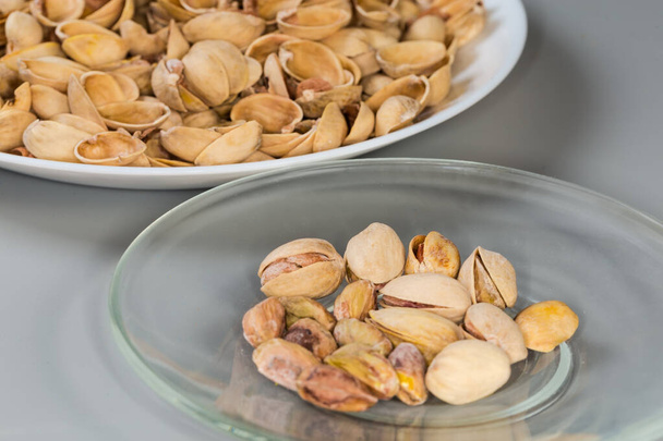 Partly peeled roasted salted pistachio nuts on the glass saucer against the empty shells on a big dish on a gray background, fragment side view in selective focus - Photo, Image