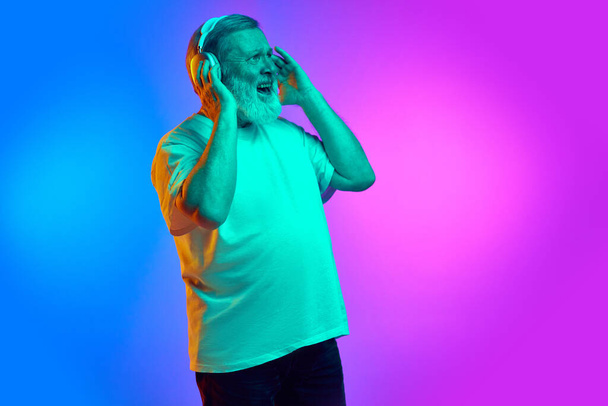 Emotional, bearded, senior man in casual clothes listening to music in headphones on gradient blue-purple background in neon light. Concept of human emotions, lifestyle, casual fashion - Foto, imagen
