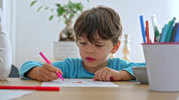 小さな男の子はカラフルなマーカーのバケツの横のテーブルで着色する宿題を楽しんでいます - 映像、動画