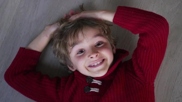 Один щасливий молодий хлопчик посміхається на камеру, лежачи на підлозі, дивлячись на камеру з блакитного кольору. Усміхнений 7-річний хлопчик з червоним светром - Кадри, відео