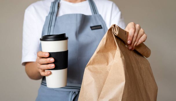 Takeaway meal in hands of waitress. Travel coffee mug and brown paper package bag with food inside.  - Photo, Image