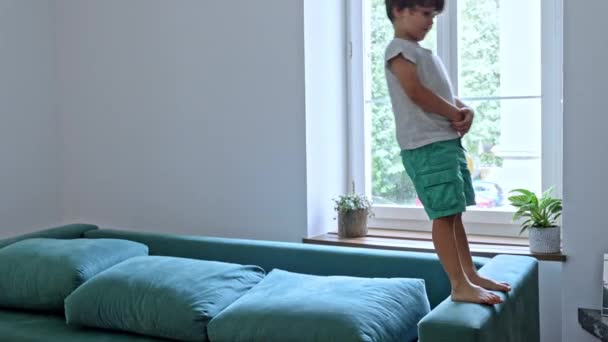 Escenas divertidas en casa: el niño pequeño sube en el brazo del sofá y cae hacia atrás en los cojines - Metraje, vídeo