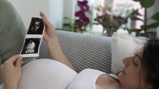 Mujer embarazada mira la imagen de ultrasonido de su bebé durante la etapa final del embarazo acostado en el sofá descansando en casa abrazando la alegría materna - Imágenes, Vídeo