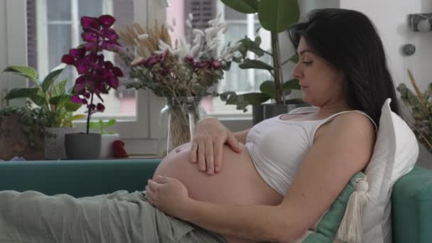 Беременная женщина отдыхает на диване и ласкает живот в предвкушении новой жизни. Профиль женщины 30-х годов во время третьего триместра беременности - Кадры, видео
