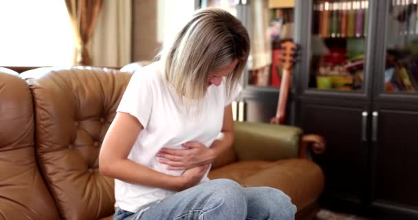 胃の痛みが激しいため、女性は手で胃をつつくソファに座っています。下痢性胃炎、リビングルームでの腸炎、人々の健康問題の概念を持つ女の子 - 映像、動画