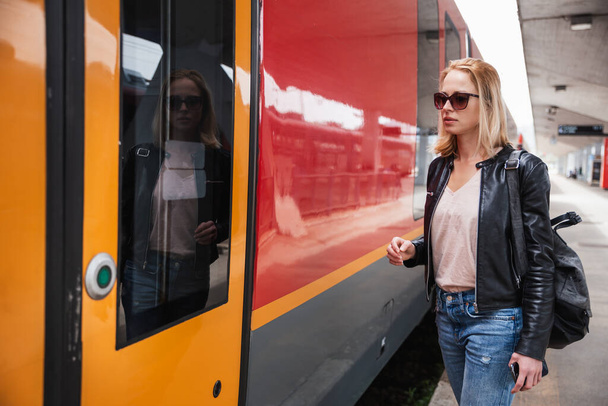 Fiatal, szőke nő farmerben, ingben és bőrdzsekiben, táskában és napszemüvegben, vörös, modern vonattal a vasútállomáson. Utazás és közlekedés - Fotó, kép