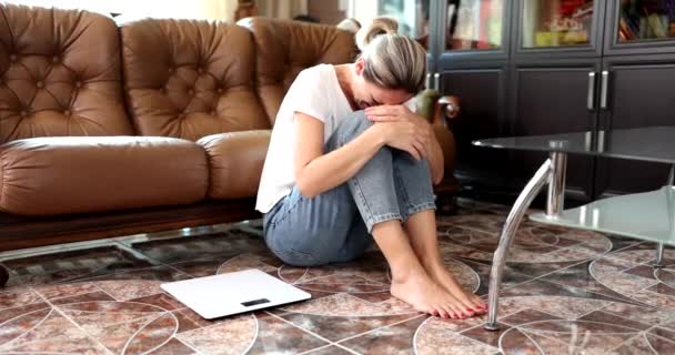 La mujer está llorando sentada en el suelo junto a escalas para medir la maasa del cuerpo. Depresión y estrés por sobrepeso - Imágenes, Vídeo