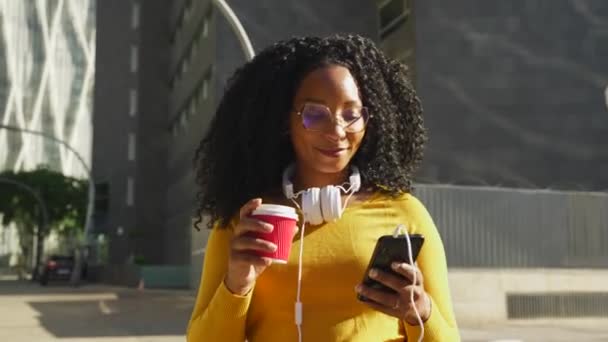 Όμορφη μαύρη γυναίκα με σγουρά μαλλιά και ακουστικά περπατάει ενώ χρησιμοποιεί το smartphone της για να συνομιλήσει με φίλους. συνδεδεμένη έννοια. - Πλάνα, βίντεο