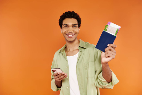 attrayant homme afro-américain joyeux tenant téléphone et passeport avec billet et souriant à la caméra - Photo, image