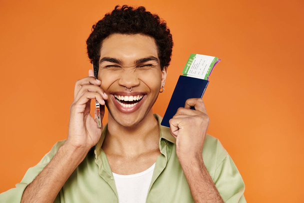 χαρούμενα ελκυστική Αφροαμερικανός άνδρας σε ζεστή ενδυμασία μιλάμε από το τηλέφωνο κρατώντας διαβατήριο με εισιτήριο - Φωτογραφία, εικόνα