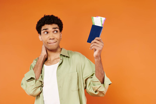 Güzel görünümlü Afrikalı Amerikalı adam turuncu arka planda bilete ve pasaporta bakıyor. - Fotoğraf, Görsel
