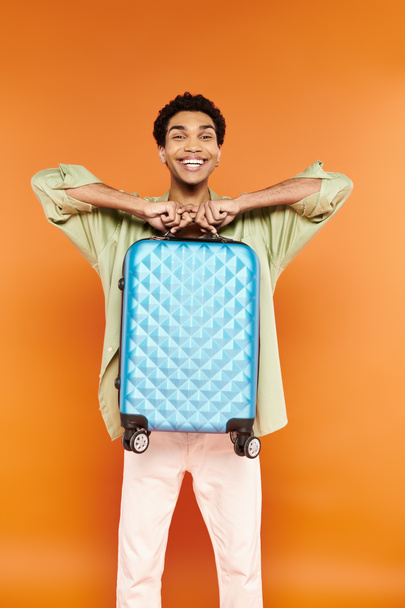 cheerful handsome african american man in casual outfit holding blue suitcase and smiling at camera - Photo, Image