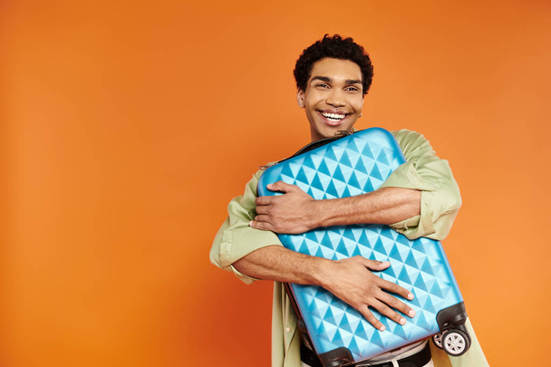 guapo y alegre hombre afroamericano con elegante atuendo abrazando su maleta azul sobre fondo naranja - Foto, imagen