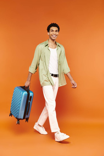 vreugdevolle aantrekkelijk Afrikaans amerikaanse man in casual outfit met blauwe koffer en glimlachen op de camera - Foto, afbeelding