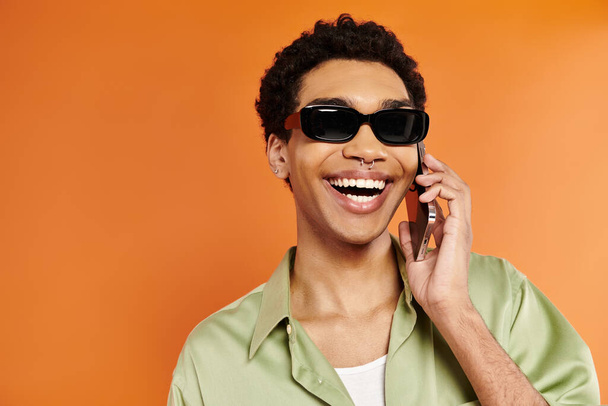 verführerisch fröhlicher afrikanisch-amerikanischer Mann mit stylischer Sonnenbrille telefoniert auf orangefarbenem Hintergrund - Foto, Bild