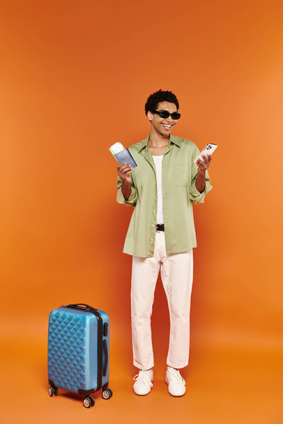radosny Afroamerykanin w okularach przeciwsłonecznych z biletem i paszportem obok swojej niebieskiej walizki - Zdjęcie, obraz