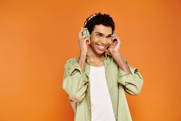 オレンジ色の背景を探しているヘッドフォンで居心地の良い服装の楽しいスタイリッシュなアフリカ系アメリカ人男性 - 写真・画像