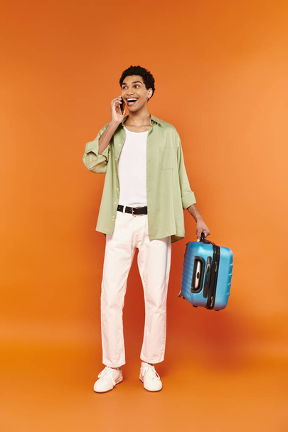 atractivo jolly africano americano hombre en acogedor traje sosteniendo maleta y hablando por teléfono móvil - Foto, imagen