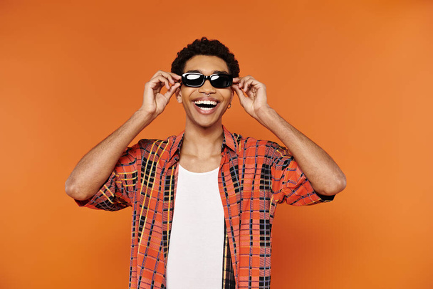 veselý mladý africký Američan muž v pulzující oblečení se slunečními brýlemi pózovat na oranžovém pozadí - Fotografie, Obrázek