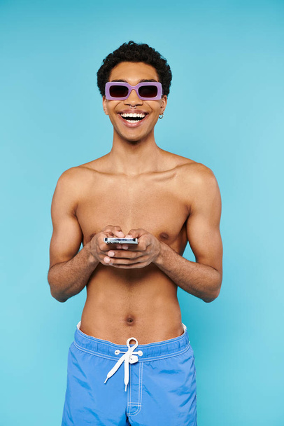 joyeux homme afro-américain avec des lunettes de soleil élégantes dans les maillots de bain tenant son smartphone - Photo, image